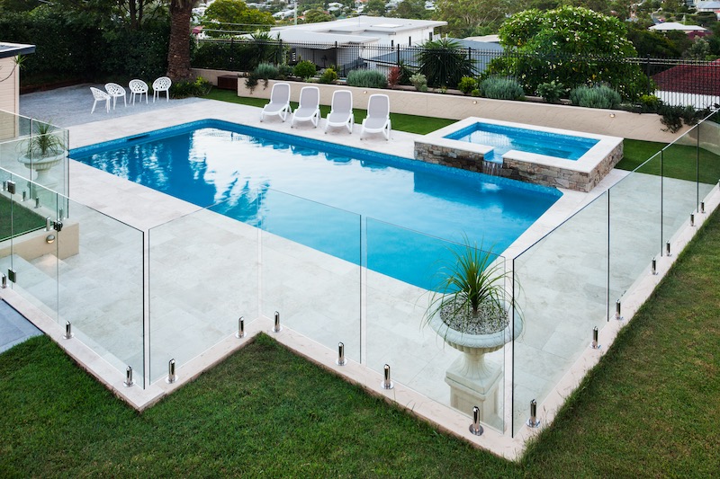 Modern swimming pool covered with glass panels beside a green lawn garden including trees and chairs
