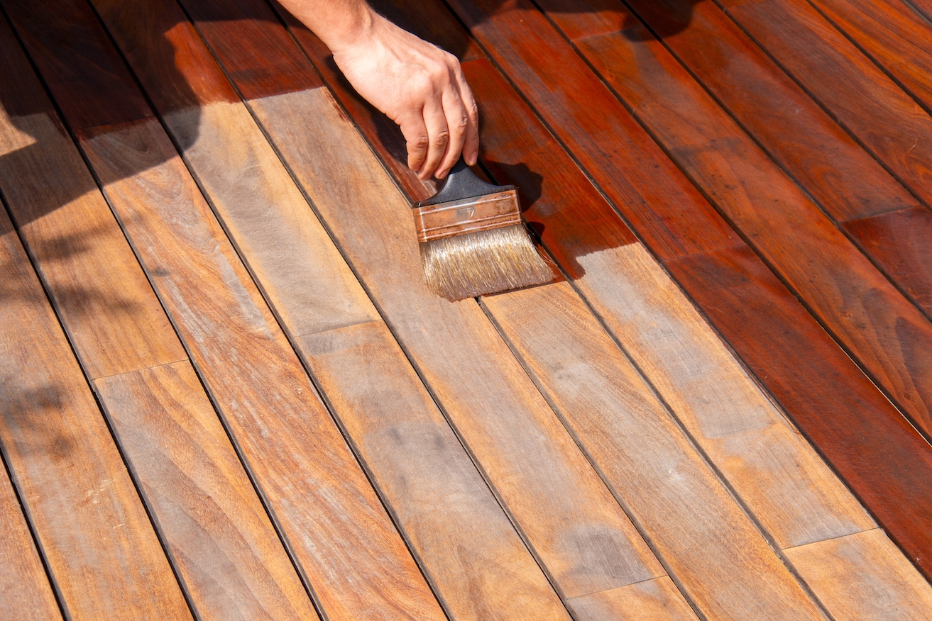 Woodworker refreshing wooden floor, worker's hand is applying decking oil on deck with a painting brush after sanding